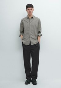 Camicia con bottoni in motivo gingham verde e bianco, maniche lunghe, abbinata a pantaloni neri e scarpe nere, posata contro uno sfondo chiaro.