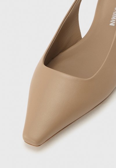 Μπεζ δερμάτινο παπούτσι μπαλαρίνα με μυτερή μύτη και σχεδιασμό με λουρί στον αστράγαλο σε ανοιχτό γκρι φόντο.