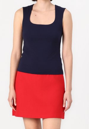 Femme portant un haut bleu marine sans manches à encolure carrée et une mini-jupe rouge vif, debout devant un fond blanc.