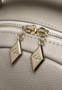 Borsa a tracolla in pelle grigia chiara, in primo piano, con zip dorate e etichette a forma di diamante con un logo stilizzato. Superficie testurizzata con dettagli.