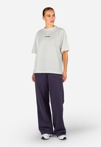 Svetlo šedé oversized tričko s krátkymi rukávmi a čiernym textovým logom, skombinované s tmavým fialovým voľným strihom nohavicami. Obe položky majú uvoľnený strih.