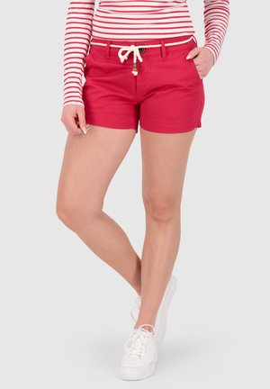 Frau trägt rote Shorts mit weißem Seilgürtel, gestreiftes Langarmshirt und weiße Turnschuhe, steht mit überschlagenem Bein.