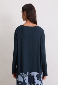 Una donna con lunghi capelli scuri indossa una maglietta lunga a maniche lunghe di un blu scuro e una gonna azzurra chiara con grandi motivi floreali scuri, rivolta di spalle rispetto alla fotocamera.