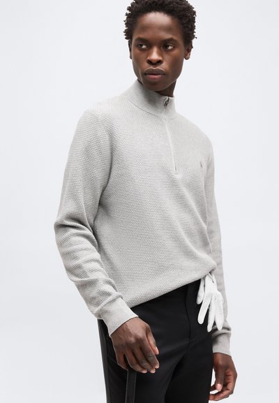 Νεαρός άνδρας που φοράει ανοιχτό γκρι υφασμένο πουλόβερ με μισό φερμουάρ και μαύρο παντελόνι, με ένα λευκό γάντι που είναι τοποθετημένο στη μέση του παντελονιού.