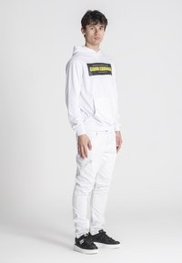 Biela mikina s čiernym a žltým logom, v kombinácii s bielymi slim-fit džínsami a čiernymi teniskami. Jednoduchý dizajn, ľahký materiál.