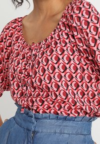 Blusa de patrones rojos y rosas con escote redondo y mangas fruncidas. El diseño presenta un motivo de ojo sobre un fondo claro.