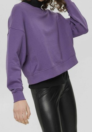 Personne portant un sweat-shirt violet ample par-dessus un haut noir et un pantalon en cuir noir moulant, un bras levé et des cheveux bouclés visibles.