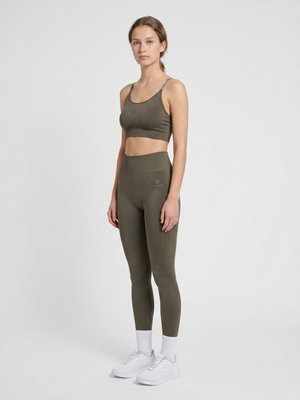 Młoda kobieta stojąca, ubrana w oliwkowo-zielony sportowy biustonosz i legginsy, białe buty sportowe i skarpetki, na jasnym, jednolitym tle.