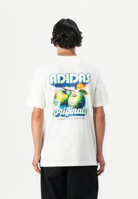 White cotton t-shirt featuring a large graphic of two green apples and the text “ADIDAS originals” in blue and green. Short sleeves, relaxed fit.