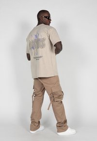 Beige grafisk t-shirt med et sommerfugl-design på ryggen, parret med brune cargo-bukser med flere lommer og stropper, hvide sneakers.