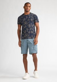 Marineblauw T-shirt met gouden bloemen- en vogelpatronen, gecombineerd met lichtblauwe cargoshorts en witte sneakers, met een ontspannen pasvorm en korte mouwen.