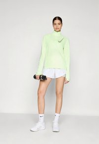 Top atlético verde-claro de manga comprida com um meio fecho de correr e um pequeno logótipo. Combinado com calções brancos e segurando halteres. Ténis brancos.