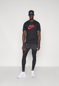 Μαύρο μπλούζα Nike με κόκκινο γραφικό "RUN", συνδυασμένο με μαύρα σορτς και μαύρους κολάν πλήρους μήκους. Λευκά αθλητικά παπούτσια και μεγάλα μπλε γυαλιά ηλίου.