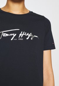 Námořnická modrá tričko vyrobené z bavlny, s kovově stříbrným logem "Tommy Hilfiger" přes hruď, s krátkými rukávy a kulatým výstřihem.