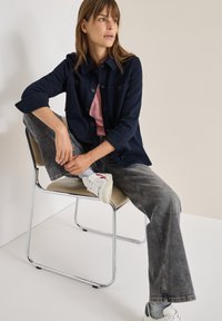 Chaqueta de botón azul marino sobre una camisa rosa, combinada con jeans grises y zapatos blancos. El modelo está sentado, con una pierna cruzada, con detalles de silla en cromo.