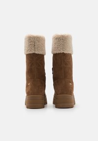 Bruna mockaskor med en fluffig beige krage, texturerad sömdetalj och en kraftig ljusbrun sula. Designade för värme och komfort.