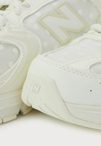White athletic shoes with mesh and leather materials, featuring a textured surface, polka dots, and a prominent, embossed logo on the sides.