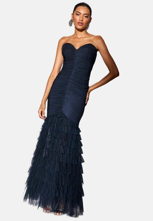 Femme portant une robe bustier bleu marine froncée avec un corsage ajusté et une jupe en tulle à volants superposés, debout, une main sur la hanche.