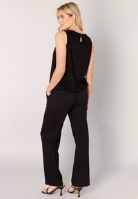 Schwarzes ärmelloses Oberteil mit einem Rückenausschnitt in Tropfenform, kombiniert mit schwarzen, weit geschnittenen Hosen und Riemchenabsätzen. Glatter Stoff, minimale Details.