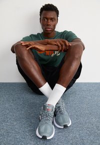 Szare buty sportowe z siateczkową cholewką, czarnymi akcentami i białą podeszwą. Model ubrany w zieloną koszulę i czarne szorty, siedzący ze skrzyżowanymi nogami.