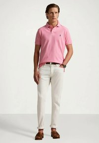 Lys rosa polo-skjorte med en blå logo, kombinert med hvite bukser og brune sko. Antrekket har en klassisk passform og korte ermer.