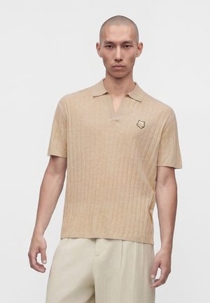 Homme à la tête rasée portant un polo côtelé beige à manches courtes avec un petit écusson animal sur la poitrine gauche et un pantalon beige clair.