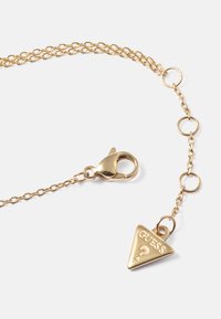 Colar de corrente dourada com um pingente triangular com "GUESS?" em relevo, fecho de lagosta e acentos de elos circulares.