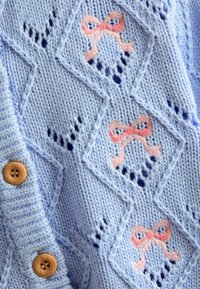 Ljusblå stickad cardigan med diamantmönster och rosa broderade rosetter. Den har en texturerad yta och knäppning med bruna knappar.