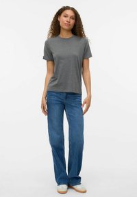 Tricou gri din bumbac cu mâneci scurte, decolteu rotund, asociat cu blugi evazați albaștri și adidași albi, stand în fața unui fundal neutru.