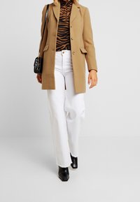 Camel uld blazer, sort og brun stribet turtleneck, hvide brede bukser, sorte ankelstøvler og en sort crossbody taske.