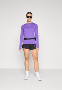 Purple long-sleeve athletic top, black shorts, and white sneakers. Features a belt with a buckle. Model wearing dark sunglasses.