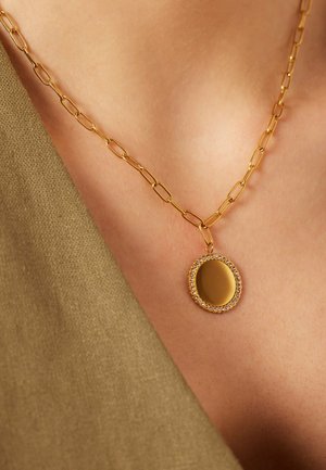 Goldene Halskette mit verlängerten Gliedern und einem runden, glatten Anhänger, der von kleinen Kristallen umrandet ist, am Hals einer Person.
