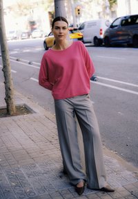 Suéter rosa de gran tamaño con mangas largas, combinado con pantalones anchos grises y zapatos marrones de punta, sobre un fondo de calle urbana.