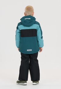 Türkisfarbene und schwarze Winterjacke mit Kapuze, bestehend aus einem farbenblockierten Design und reflektierenden Akzenten. Getragen mit schwarzen Hosen und grünen Schuhen.