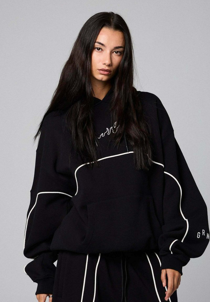 Sudadera negra de gran tamaño con detalles y logotipo blancos. Cuenta con un bolsillo frontal y capucha con cordón. Textura suave y diseño casual.