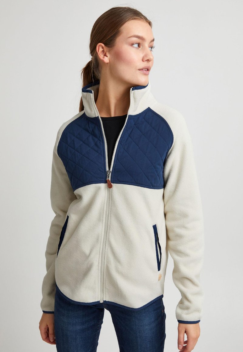 Vilnas jaka krēmkrāsā ar tumši zilu kviltētu paneli. Aprīkota ar augstu apkakli, rāvējslēdzēja aizdari un sānu kabatām ar tumši ziliem akcentiem.