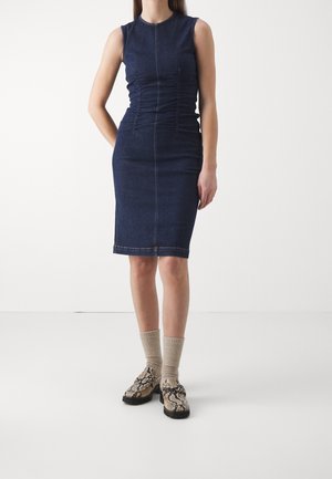 Robe sans manches en denim foncé avec des détails froncés sur les côtés, silhouette ajustée et ourlet à hauteur du genou. Associée à des chaussures à motifs et des chaussettes côtelées.