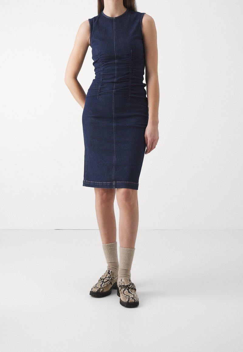 Tmavě modré džínové šaty bez rukávů s nabíranými bočními detaily, přiléhavým střihem a délku po kolena. V kombinaci s vzorovanými botami a žebrovanými ponožkami.