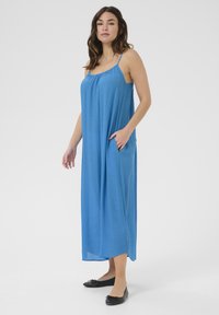 Vestido maxi azul sin mangas con un corte relajado, con bolsillos y un escote fruncido. Llevado con unas ballerinas negras.