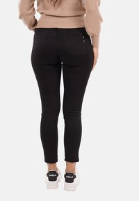 Donna che indossa jeans skinny neri e sneakers bianche, in piedi con le mani rilassate lungo i fianchi su uno sfondo semplice.