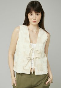 Gilet sans manches beige avec des motifs carrés texturés, une fermeture à nouer à la taille et un tissu doux et léger. Porté par-dessus un haut blanc.