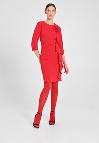 Abito rosso aderente con maniche a tre quarti, caratterizzato da un volant laterale e un'applicazione a forma di rosa, abbinato a collant rossi e sandali neri con tacco alto.