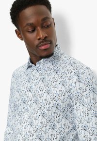 Mann trägt ein weißes Hemd mit blauen und braunen Blumenmustern, schaut nach unten und zur Seite vor einem weißen Hintergrund.