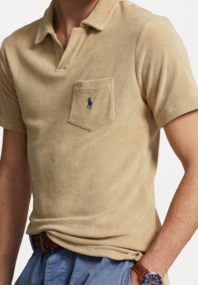 Polo en khaki avec tissu texturé, manches courtes et poche sur la poitrine. Présente un logo brodé bleu sur la poche.