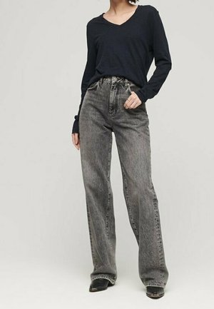 Person iført højtaljede, vidde grå jeans og en langærmet sort V-hals top, stående med den ene hånd i lommen.