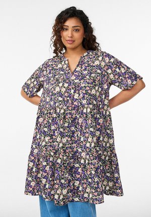 Kortsuga naine, kes kannab lahtist lillaka mustriga kleiti ja siniseid džeansse, seisab valge tausta ees käed puusal.