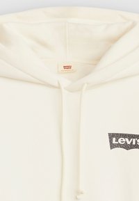 Kremo spalvos su kapuce džemperis su tekstūruota paviršiumi, su raišteliais ir juodu leopardų rašto Levi's logotipu ant krūtinės.