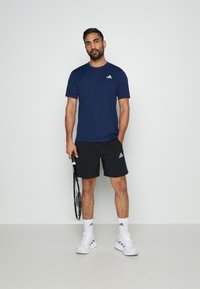 Camiseta atlética azul y pantalones cortos negros, ambos con el logo de Adidas. El modelo sostiene una raqueta de tenis. Zapatillas y calcetines blancos completan el atuendo.