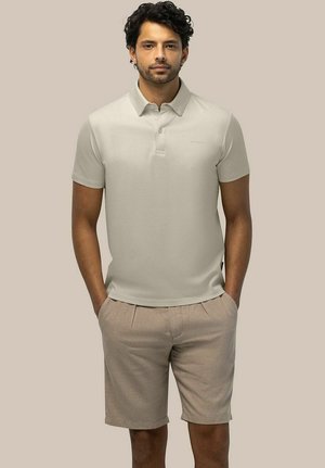 Man with curly hair wearing a light beige short-sleeve polo shirt and beige shorts standing with hands in pockets against plain background.