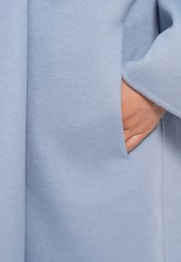 Abrigo de mezcla de lana azul claro con una textura suave y bolsillo lateral, que presenta un corte relajado y detalles de diseño minimalistas.